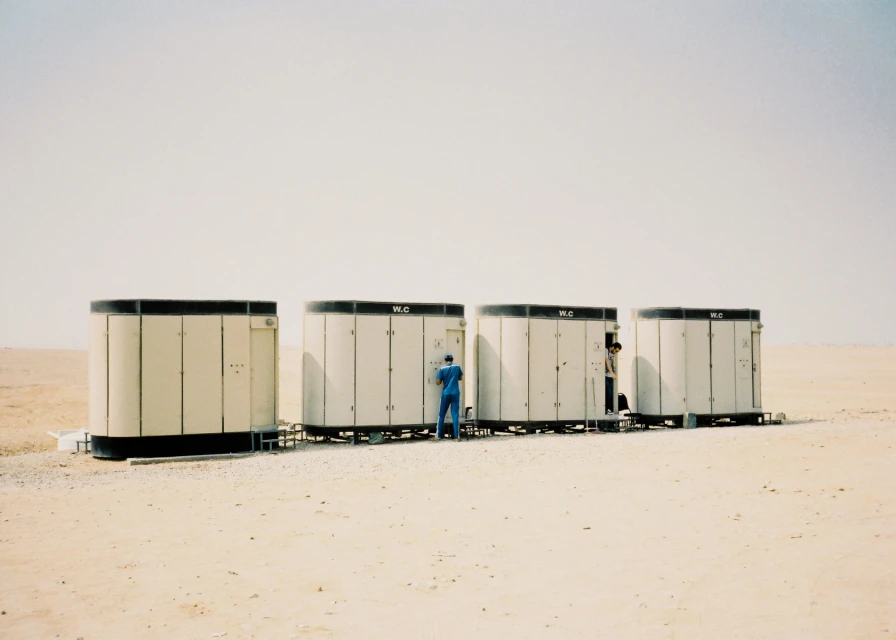 Four portable restrooms stand in the desert landscape, each with a 'WC' sign, and two men are interacting with the facilities.