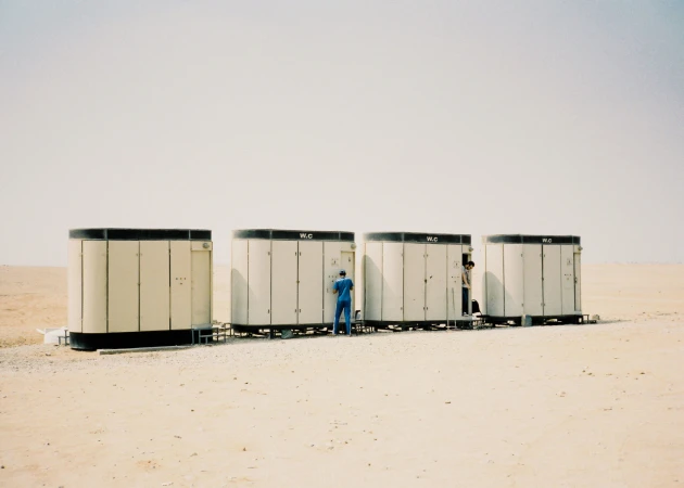 Four portable restrooms stand in the desert landscape, each with a 'WC' sign, and two men are interacting with the facilities.
