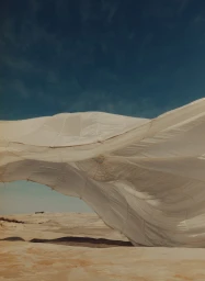 Large white fabric structure blowing in the wind above a sandy desert landscape under a blue sky.