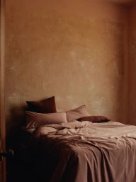 A neatly made bed with mauve linens and dark brown pillows against a textured terracotta wall, photographed by Daniel Goode.