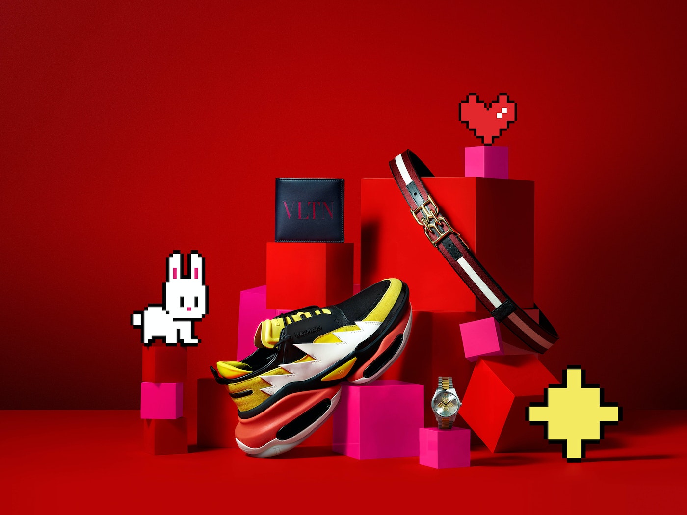 Still life product shot featuring a colorful pair of sneakers, a wallet, a belt, and a watch on pink and red blocks against a red backdrop.