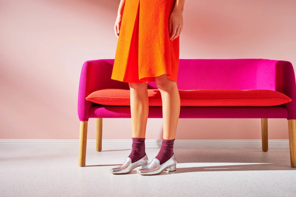 Woman in orange dress, metallic silver loafers, and burgundy socks standing near a pink sofa against a light pink backdrop.