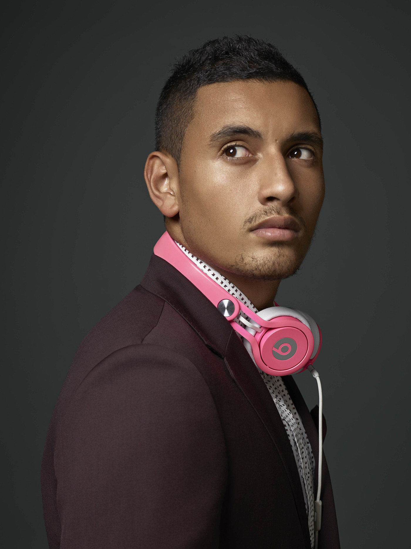 Close-up studio portrait of adult male glancing left; wearing burgundy suit jacket, patterned shirt, and pink headphones around his neck.