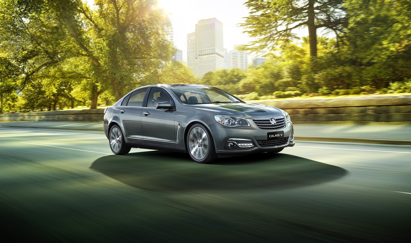 Grey Holden Calais V car in motion on a road with green trees and an urban skyline in the background on a sunny day.