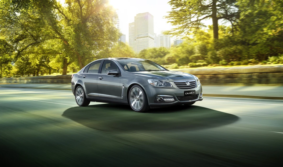 Grey Holden Calais V car in motion on a road with green trees and an urban skyline in the background on a sunny day.