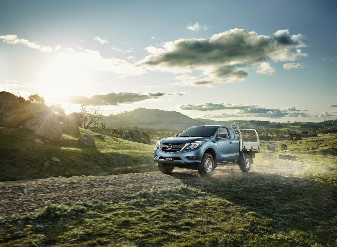 Blue Mazda BT-50 pickup truck drives on a dirt road through a grassy field with hills, trees, and a cloudy sky background.