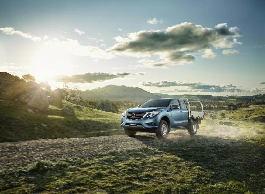Blue Mazda BT-50 pickup truck drives on a dirt road through a grassy field with hills, trees, and a cloudy sky background.