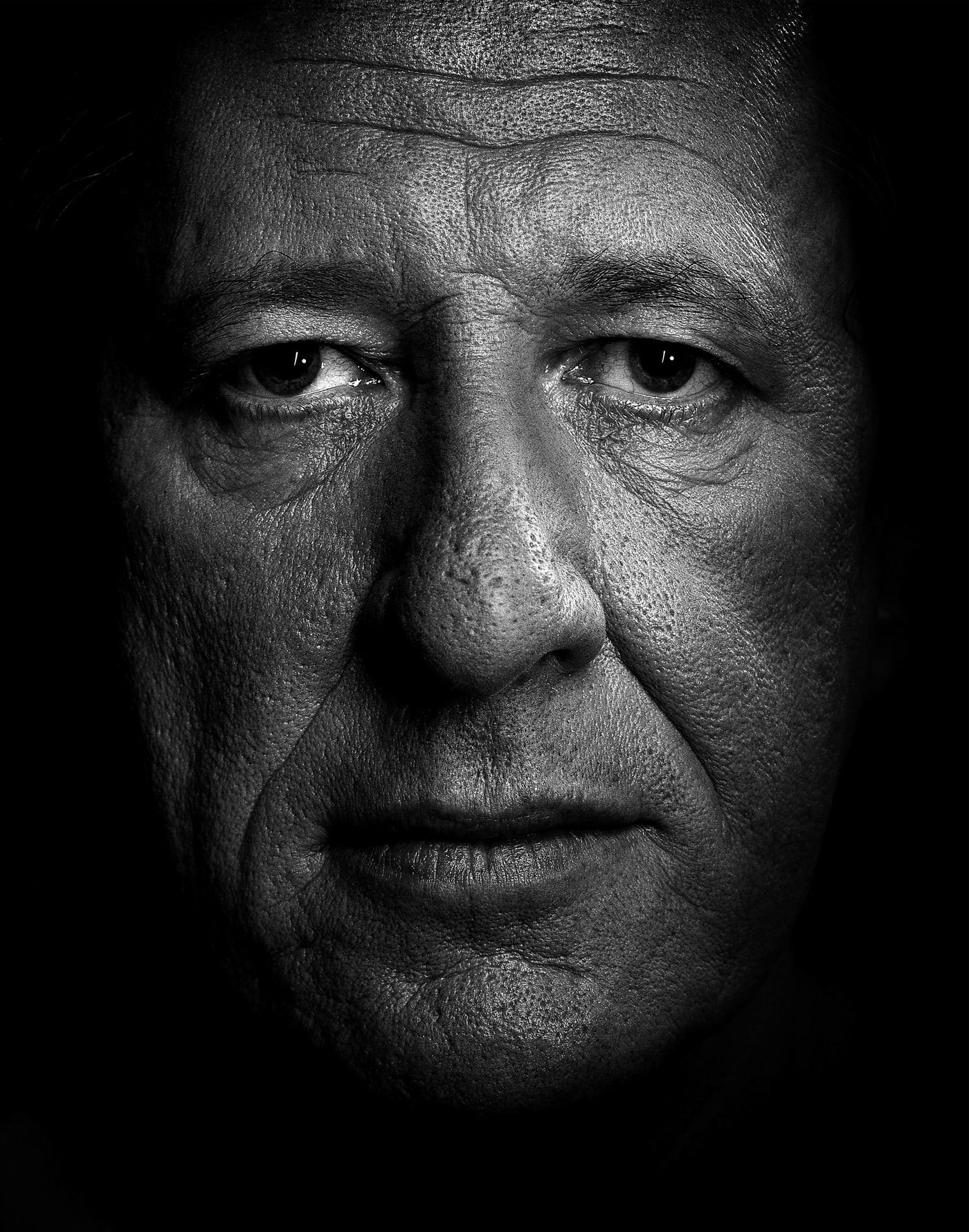 Monochrome close-up portrait of a mature adult male, showing facial details, skin texture, and low-key lighting that emphasizes shadows.