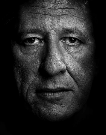 Monochrome close-up portrait of a mature adult male, showing facial details, skin texture, and low-key lighting that emphasizes shadows.