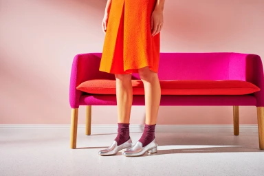 Model wearing a bright orange skirt, sparkling socks, and silver loafers standing before a pink sofa
