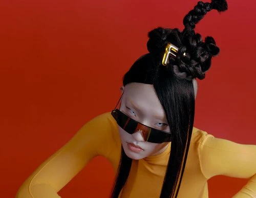 Model with avant-garde braided hairstyle wearing yellow top and sunglasses against red background