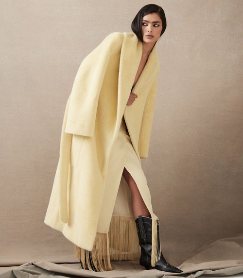 Fashionable woman poses in studio, wearing a long yellow coat, skirt with fringe, and cowboy boots against a neutral backdrop.