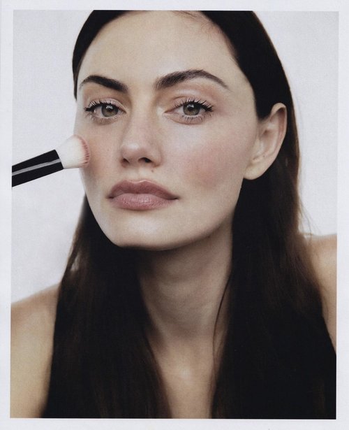 Close-up shot of an adult woman with long dark hair applying blush to her cheek with a makeup brush in a white photo studio.
