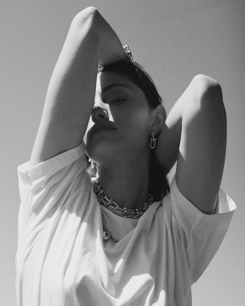 Black and white portrait of a woman posing in direct sunlight wearing multiple chain necklaces, hoop earrings, and a T-shirt.