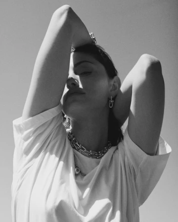 Black and white portrait of a woman posing in direct sunlight wearing multiple chain necklaces, hoop earrings, and a T-shirt.