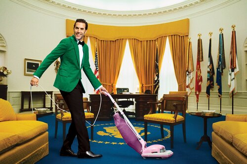 Man in a green blazer vacuuming a rug in an oval office replica with yellow chairs and sofas, plus American flags and a desk visible.