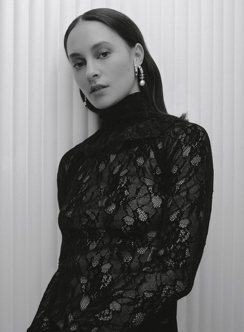 Portrait of an adult female wearing a black lace turtleneck top with pearl earrings, posing indoors against a white vertically striped wall.