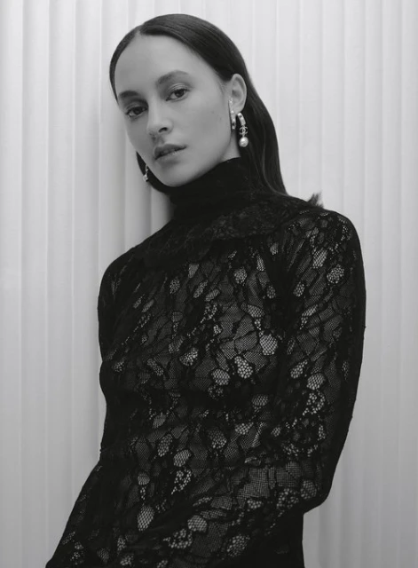 Portrait of an adult female wearing a black lace turtleneck top with pearl earrings, posing indoors against a white vertically striped wall.