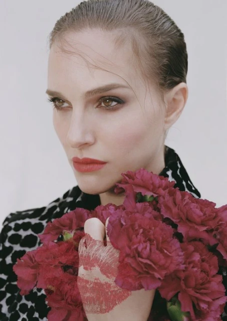 Portrait of a woman holding red carnations and wearing a black-and-white patterned jacket against a plain white background.