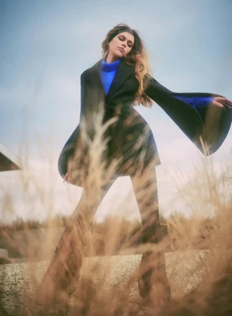 Low-angle shot of a woman in a black coat and blue sweater standing behind tall dry grass.