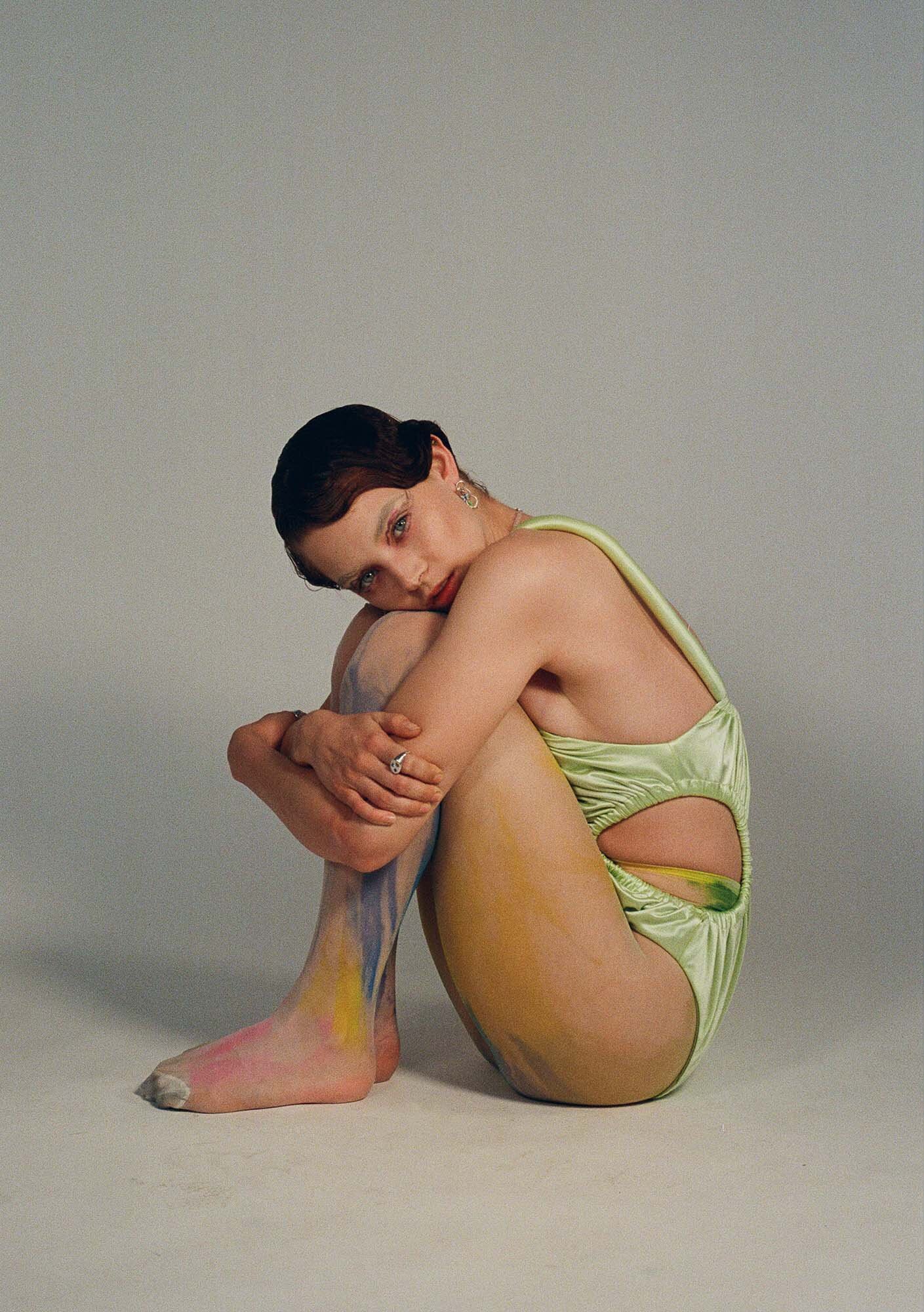 Woman in a light green leotard sits and hugs her knees in a studio with a neutral background, showcasing a contemporary fashion image.
