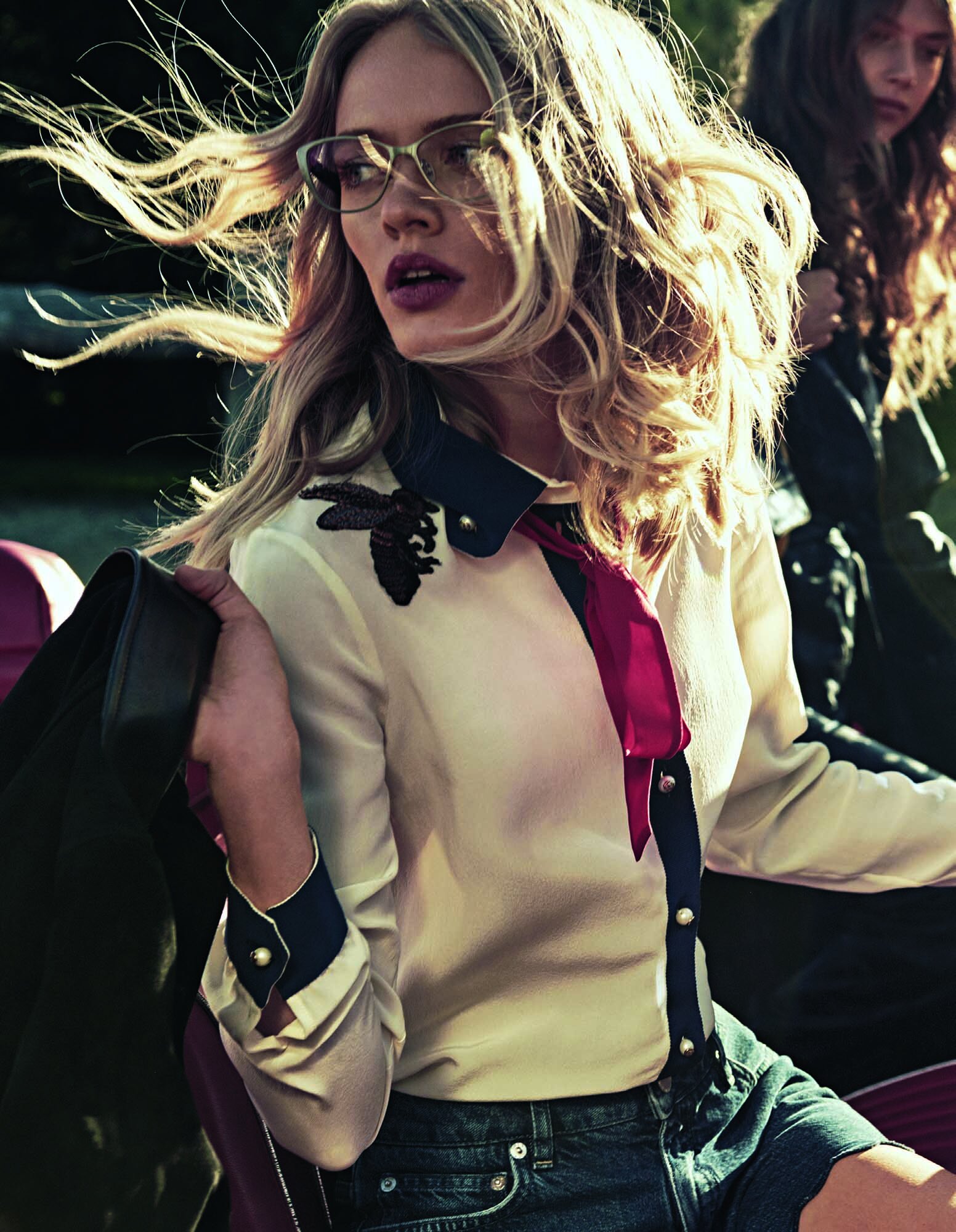 Two women in outdoor fashion shot: one blonde with glasses, wearing a collared shirt and denim shorts, and another woman behind.