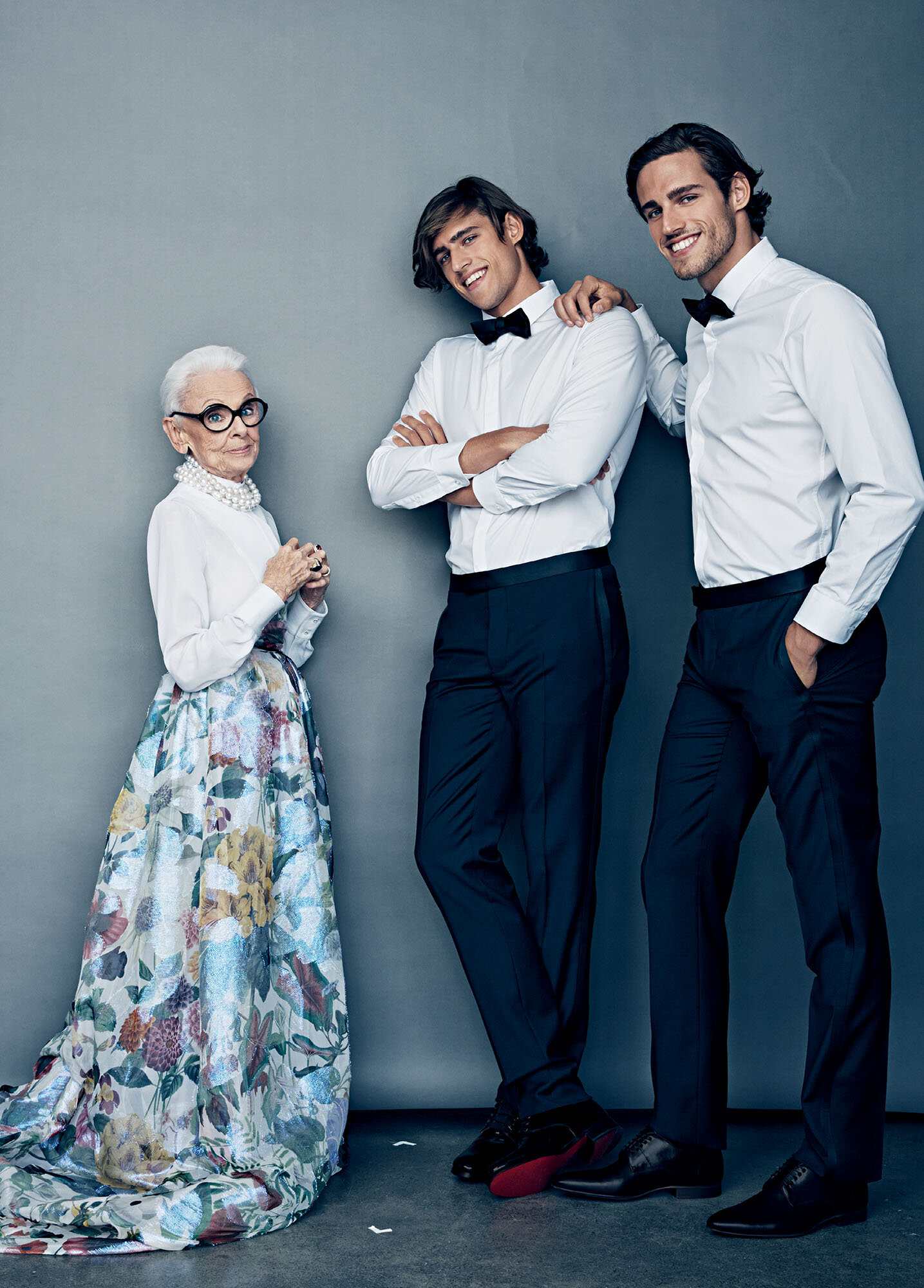 Elderly woman in floral skirt poses with two young men in tuxedos against a gray backdrop in a studio setting. Formal wear fashion.