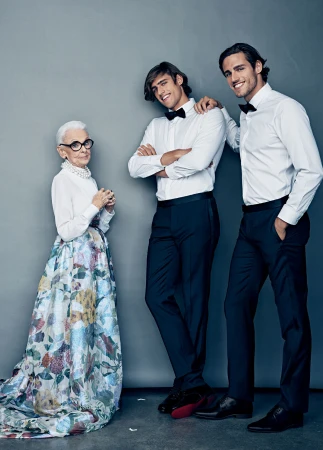 Elderly woman in floral skirt poses with two young men in tuxedos against a gray backdrop in a studio setting. Formal wear fashion.