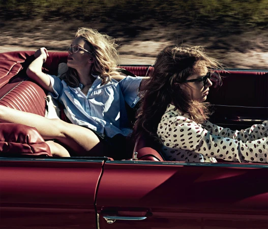 Two young adult women ride in a red convertible car on a bright day. One woman drives; the other relaxes in the passenger seat.
