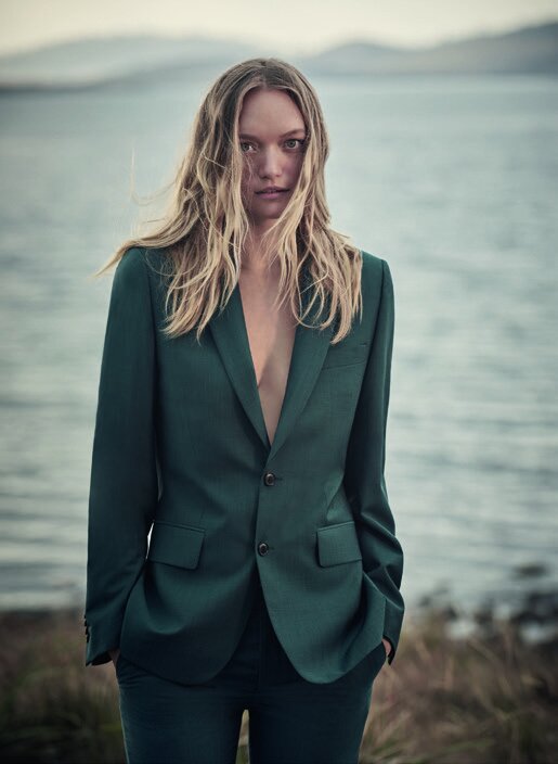 Adult female model in a green suit poses outdoors with wavy blonde hair, portrait orientation, against a blue water and shoreline background.