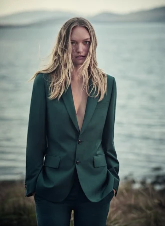 Adult female model in a green suit poses outdoors with wavy blonde hair, portrait orientation, against a blue water and shoreline background.