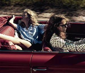 Two women riding in a red vintage convertible car with blurred roadside scenery passing by in the background.