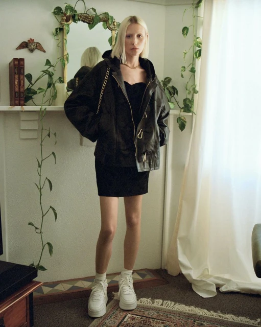 Full-length shot of a young, fair-skinned woman with blonde hair in a black dress and jacket, standing indoors with neutral décor.