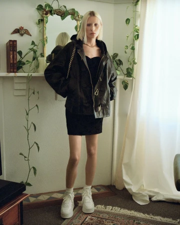 Blonde woman wearing an oversized dark leather jacket and white sneakers standing in a bright living room.