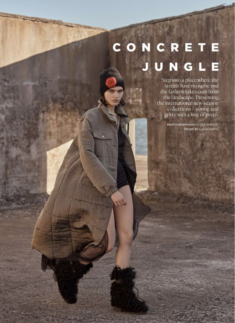 Model in a long quilted olive coat, black beanie with red flower, and furry boots walking past concrete ruins.