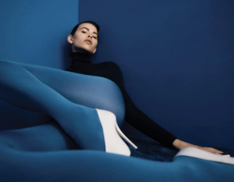 A woman with dark hair and a black turtleneck sweater poses against a blue wall, wearing blue tights and white high-heeled shoes.