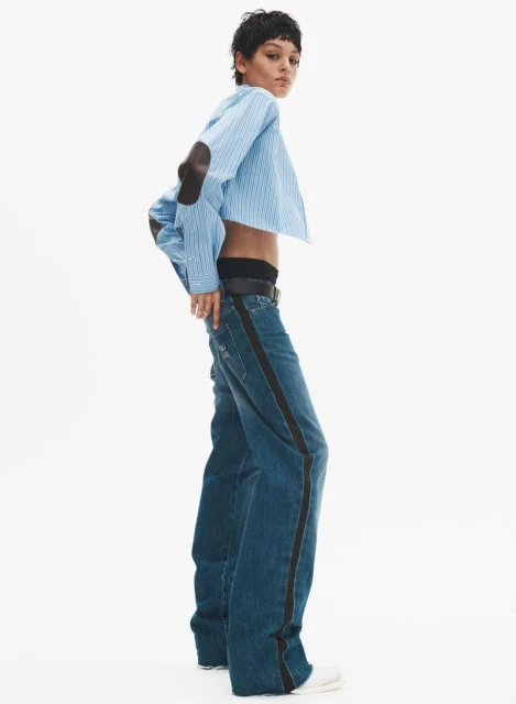 An adult woman poses in a blue striped shirt and denim jeans, styled in a cropped fashion, set against a bright white backdrop.