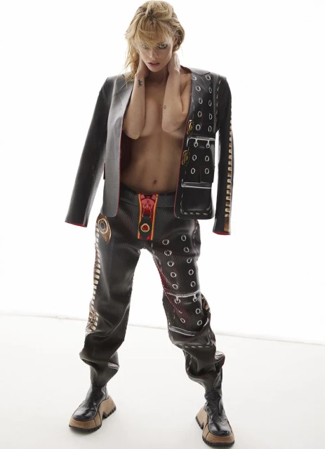 Blonde-haired person wearing a black leather jacket and pants, posing in a white studio. Fashion editorial showcasing alternative styles.