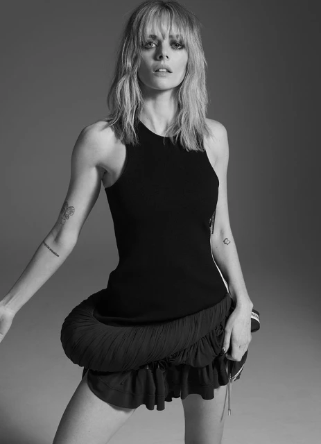 Black and white portrait of a woman with blonde hair, wearing a black top and a dark ruffled skirt, posing in a photo studio.