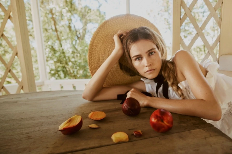 Blonde woman in straw hat rests at wooden table with peaches and nectarines in outdoor setting with natural lighting and neutral palette.