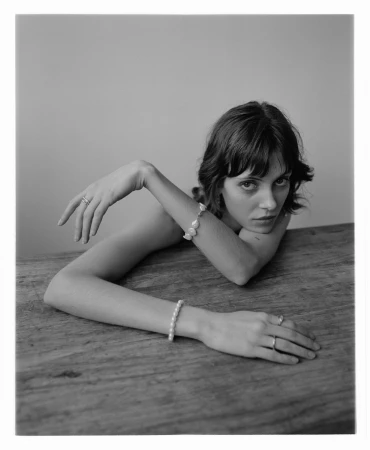 Monochrome portrait of a woman resting on a wooden table, wearing pearl bracelets and rings with make-up by Peter Beard.