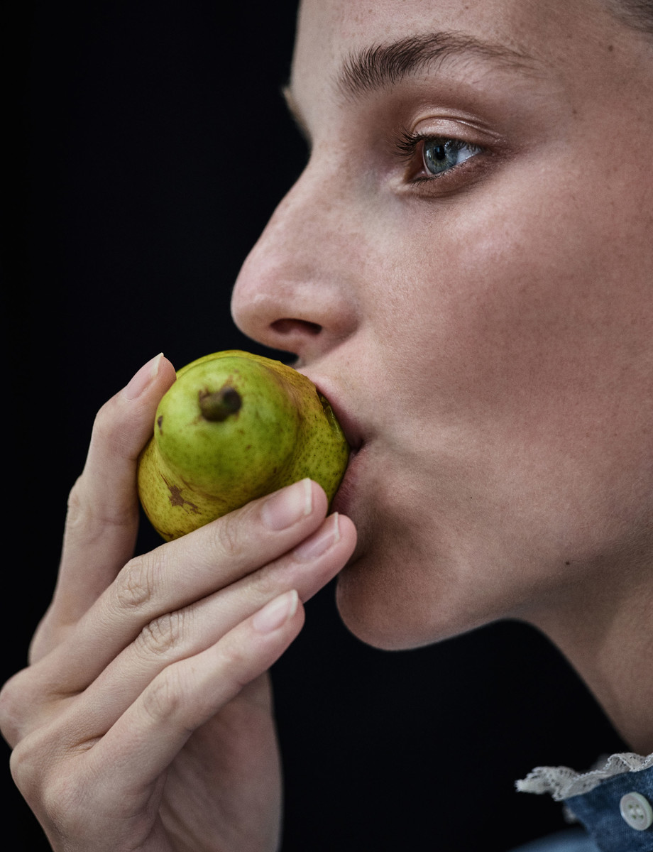 Close-up profile of a woman holding a small green pear near her lips against a solid dark background