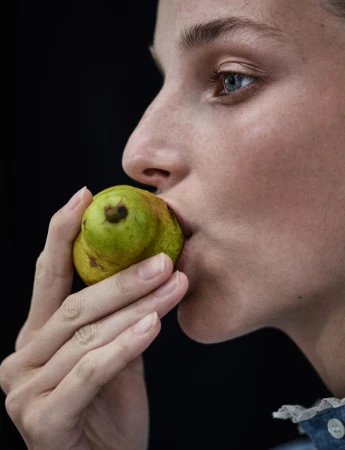 Close-up profile of a woman holding a small green pear near her lips against a solid dark background