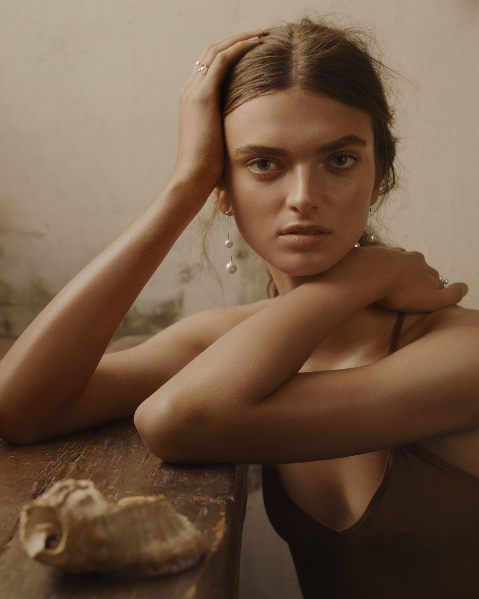 Portrait of a young woman with brown hair, wearing pearl earrings, with her hands on her head, posing in an indoor setting.