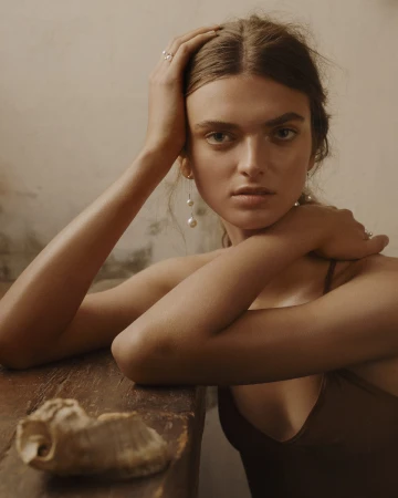 Portrait of a young woman with brown hair, wearing pearl earrings, with her hands on her head, posing in an indoor setting.