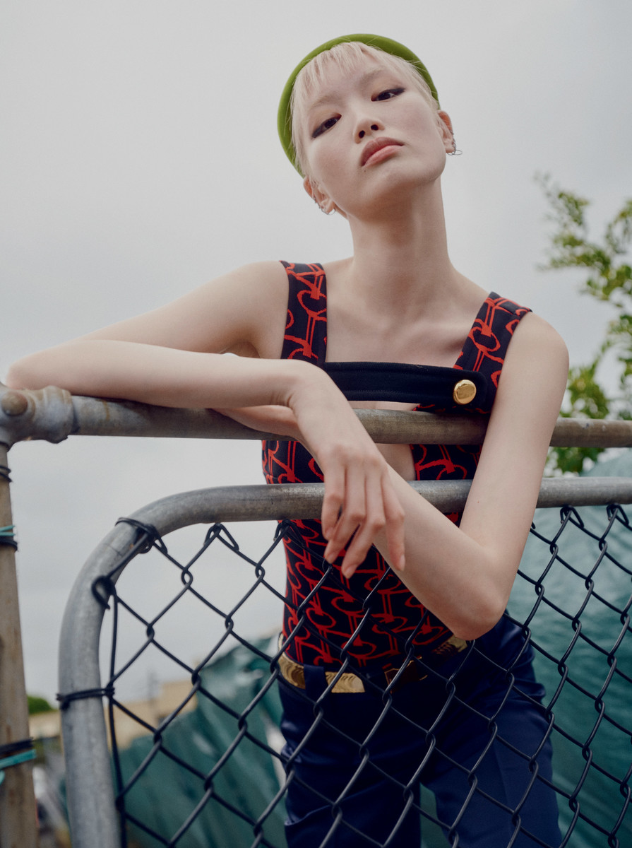 A blonde woman poses in a patterned red and black tank top, dark pants, and a green beanie hat, leaning on a gray metal fence.