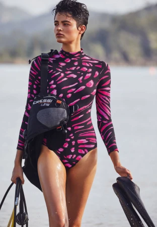 Woman in a pink and black wetsuit stands on a sandy shore with diving gear, including flippers and a Cressi dive bag, on a sunny day.