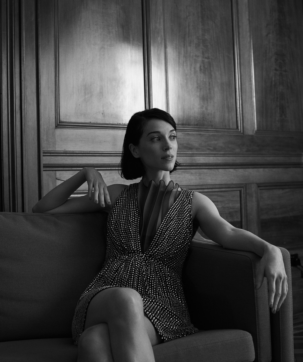Monochrome portrait of a woman with dark bobbed hair seated on a sofa wearing a patterned dress against wooden paneling.