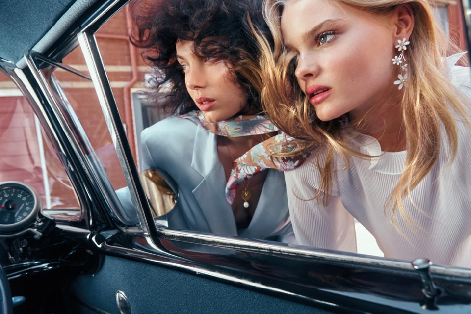 Two women inside a vintage car. One has curly dark hair and a light blue blazer, the other has blond hair and a white blouse.