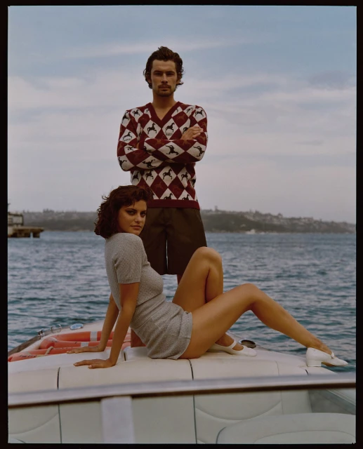 Stylish couple poses on a boat with seascape backdrop. The woman sits in a grey knit dress while the man stands behind in a patterned sweater.